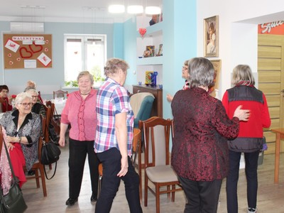 Zdjęcie przedstawia seniorki tańczące wokół krzeseł podczas zabawy w poszukiwanie fantów.