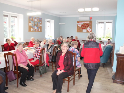 Na zdjęciu znajdują się seniorki biorące udział w zabawie w „gorące krzesła”. Widać również osoby siedzące przy stołach. W tle gazetka ścienna o tematyce walentynkowej.