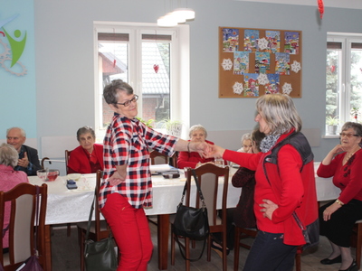 Zdjęcie przedstawia dwie tańczące seniorki. W tle osoby starsze siedzące przy stole.