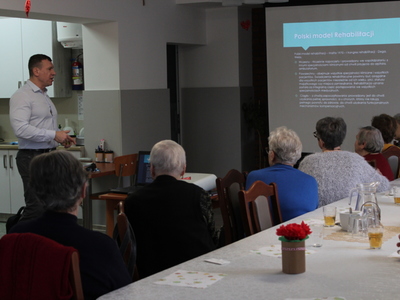 Zdjęcie przedstawia wykładowcę tłumaczącego seniorom kwestie dotyczące zajęć rehabilitacyjnych.