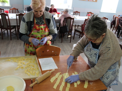 Na zdjęciu jedna z pań kroi ciasto na faworki, a druga formuje ciastka. W tle seniorzy siedzący przy stole.