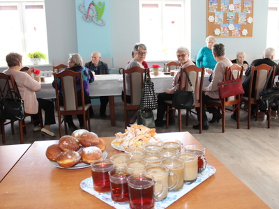 Na pierwszym planie zdjęcia: kawa, herbata, faworki i pączki. W tle seniorzy siedzący przy stołach.
