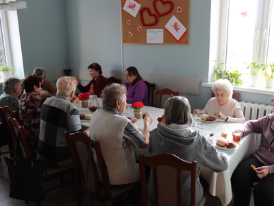 Zdjęcie przedstawia seniorki siedzące przy stole. Wszyscy delektują się samodzielnie wypieczonymi chruścikami oraz pączkami.