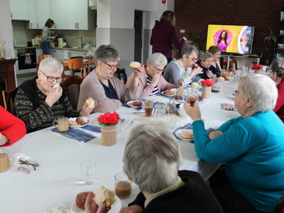 Na fotografii seniorzy jedzą pączki z okazji tłustego czwartku. W tle pracownia kulinarna.