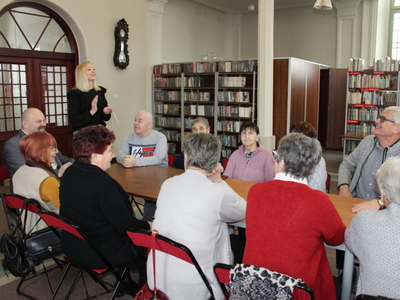 Fotografia wykonana w Miejsko – Gminnej Bibliotece Publicznej w Sompolnie. Przedstawia uczestników wykładu siedzących przy wspólnym stole oraz panią dyrektor biblioteki przedstawiającą wykładowcę.
