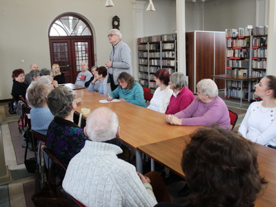 Zdjęcie wykonane we wnętrzu biblioteki. Widać na nim mężczyznę stojącego za stołem i przedstawiającego się słuchaczom wykładu.