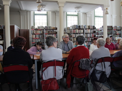 Fotografia  wykonana we wnętrzu biblioteki. Przedstawia seniorów siedzących przy stole i słuchających wykładu na temat chorób serca.