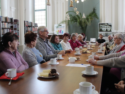 Fotografia przedstawia grupę seniorów siedzącą przy wspólnym stole wraz z wykładowcą w bibliotece.