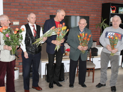 Zdjęcie wykonane w sali głównej Domu Dziennego Pobytu Senior+ w Sompolnie. Widać na nim pięciu mężczyzn stojących z kwiatami dla kobiet w dniu ich święta.