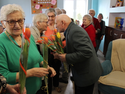 Zdjęcie przedstawia grupę seniorek oraz mężczyznę – jednego z uczestników DDP składającego życzenia jednej z kobiet.