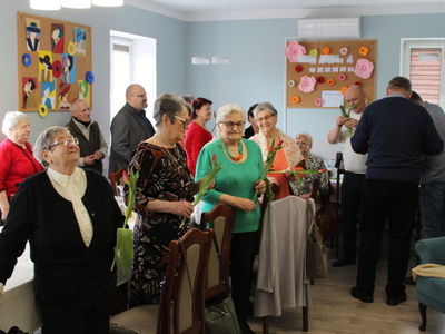 Na fotografii grupa seniorek oraz mężczyźni wręczający im kwiaty.