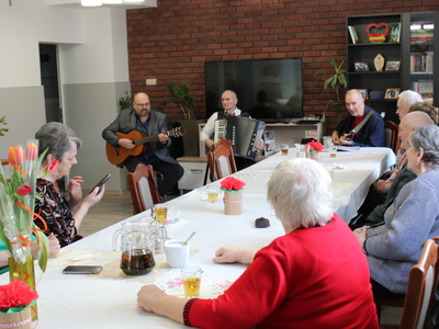 Zdjęcie przedstawia dwóch mężczyzn grających na gitarach i jednego na akordeonie oraz grupę seniorek siedzących przy stole.