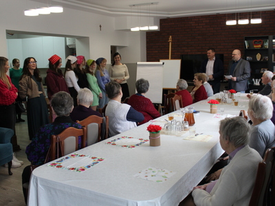 Na zdjęciu seniorzy siedzący przy stole oraz grupa młodzieży wraz z opiekunami stojący na tle pracowni kulinarnej. W tle burmistrz miasta Sompolna oraz dyrektor mgops w Sompolnie.