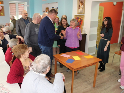 Na zdjęciu widać dwa rzędy uczestników zabawy w kolory oraz seniorów siedzących przy stole.