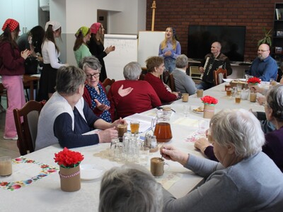 Fotografia przedstawia grupę seniorów siedzących przy stole, dwóch mężczyzn grających na instrumentach oraz dziewczynę śpiewającą razem z nimi. Po prawej stronie grupa młodzieży oklaskująca występ.