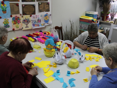 Zdjęcie przedstawia cztery seniorki siedzące przy stole w pracowni artystycznej i składające elementy do kurczaka, wykonywanego metodą origami modułowego.