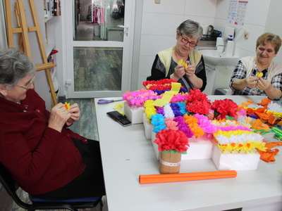 Fotografia przedstawia trzy seniorki siedzące przy stole w pracowni plastycznej i układające kwiaty z bibuły.
