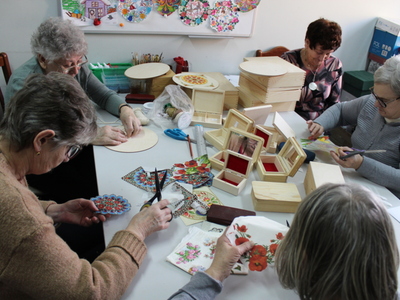 Zdjęcie prezentuje pięć seniorek siedzących przy stole w pracowni plastycznej, które wycinają wiosenne motywy do ozdabiania pudełek metodą decoupage.