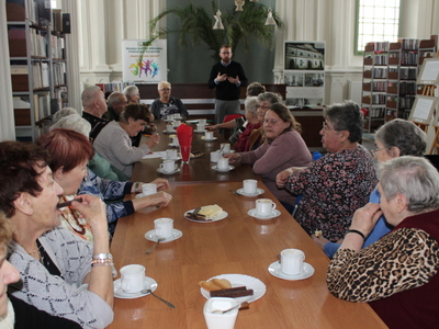 Fotografia wykonana w Miejsko – Gminnej Bibliotece Publicznej w Sompolnie. Przedstawia seniorów siedzących przy stole. W tle wykładowca opowiadający o możliwościach internetu.