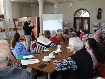 Zdjęcie przedstawia seniorów siedzących przy stole. Z lewej strony stoi wykładowca przedstawiający prezentację na temat Internetu.