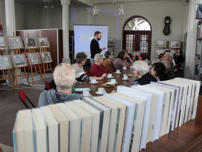 Na zdjęciu znajdują się seniorzy siedzący przy stole w bibliotece oraz mężczyzna prowadzący wykład. Z lewej strony sztalugi z plakatami dotyczącymi powstania styczniowego.