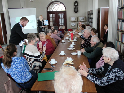 Zdjęcie przedstawia wykładowcę wręczającego upominki seniorom siedzącym przy stole. W tle pani dyrektor biblioteki prezentująca podziękowanie dla uczestników wykładów.