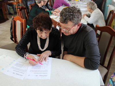 Zdjęcie wykonane w sali głównej Domu Dziennego Pobytu Senior+ w Sompolnie. Przedstawia wolontariusza, który pomaga jednej z seniorek rozwiązywać zadanie poprawiające pamięć .