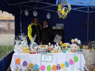 Zdjęcie przedstawia stoisko Domu Dziennego Pobytu zaprezentowane podczas kiermaszu Wielkanocnego w dniu 1 kwietnia 2023r. W tle dwie pracownice DDP.