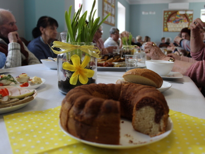 1	Zdjęcie stołu wielkanocnego: na pierwszym planie dekoracje i potrawy, a w tle seniorzy.
