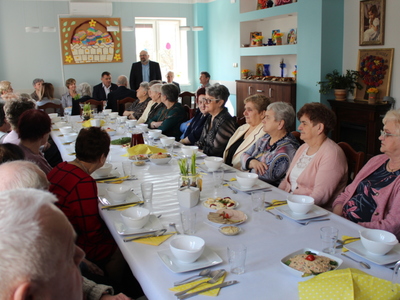 Zdjęcie wykonane w Domu Dziennego Pobytu Senior+ w Sompolnie. Przedstawia wszystkich uczestników siedzących przy stole. W tle widać zaproszonych gości oraz dyrektora MGOPS, który w pozycji stojącej przemawia do zgromadzonych.