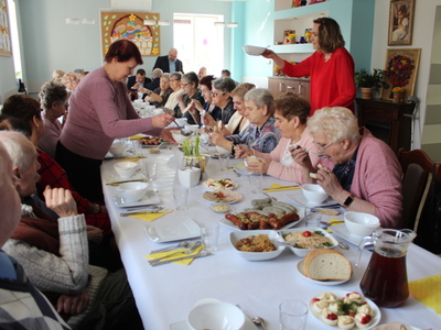 Zdjęcie przedstawia seniorów siedzących przy stole i spożywających śniadanie wielkanocne. Po prawej stronie jedna z pań nalewa żurek, a po lewej stronie opiekunka osób starszych donosi miskę z jedzeniem.