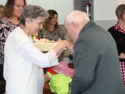 Zdjęcie przedstawia jednego z seniorów składającego życzenia jubilatce. W tle zaproszeni goście.