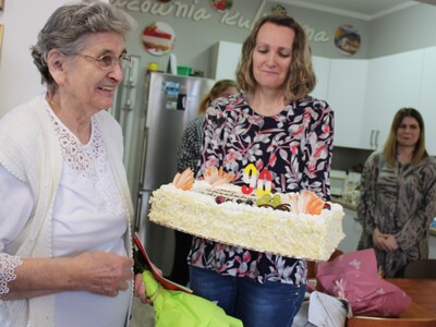 Na fotografii uśmiechnięta jubilatka. Obok niej jedna z pracownic DDP trzymająca okazały tort. W tle pracownia kulinarna.