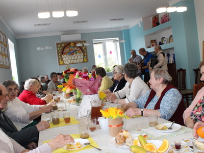 Zdjęcie przedstawia wszystkich uczestników urodzin siedzących przy stołach. W tle trzej mężczyźni grający na instrumentach: dwóch gitarach i akordeonie.
