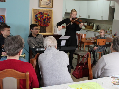 Zdjęcie wykonane w sali głównej Domu Dziennego Pobytu. Przedstawia seniorów siedzących przy stole oraz czworo dzieci: dwóch chłopców i dwie dziewczynki. Jedna z  dziewczyn gra na skrzypcach. W tle pracownia kulinarna.