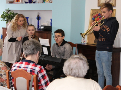 Zdjęcie przedstawia kobietę pochylającą się nad uczennicą, chłopca przy keyboardzie oraz chłopca grającego na trąbce.