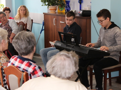 Zdjęcie przedstawia chłopca grającego na keyboardzie. W tle opiekun dzieci oraz grupa seniorów siedzących przy stole.