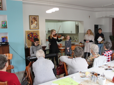 Na zdjęciu wszyscy uczniowie wraz z nauczycielką, którzy przyszli do DDP zaprezentować koncert wiosenny, oraz seniorzy siedzący przy stole wsłuchani w grę na instrumentach.