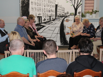 Zdjęcie wykonane w sali głównej domu dziennego pobytu senior+ w Sompolnie. Przedstawia siedmioro seniorów siedzących na ławkach i grających w sztuce pt: „Sąsiedztwo”. Przed seniorami widać publiczność siedzącą na krzesłach. W tle scenografia do sztuki przedstawiająca namalowane czarną farbą bloki, chodnik, drzewo.