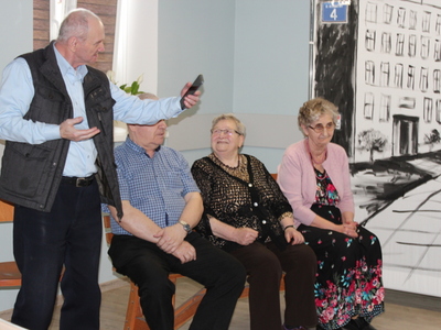 Zdjęcie przedstawia trójkę seniorów – aktorów, siedzących na ławce podczas przedstawienia oraz starszego mężczyznę stojącego obok ławki i pokazującego telefon komórkowy.