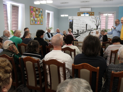 Zdjęcie przedstawia widownię oglądającą spektakl. W tle seniorzy – aktorzy i scenografia. Po prawej stronie dwaj mężczyźni opowiadający o sztuce.