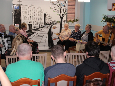Zdjęcie przedstawia seniorów siedzących na ławkach, śpiewających piosenkę na zakończenie spektaklu. Wśród nich mężczyzna grający na akordeonie oraz drugi – na gitarze.