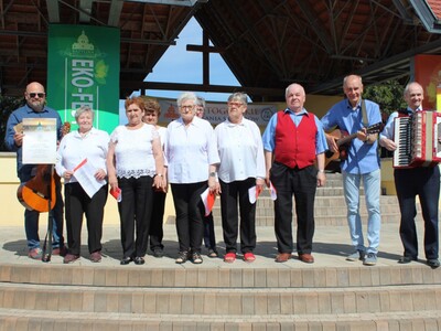 Fotografia wykonana na plenerowej scenie w Świętej Górze. Na zdjęciu seniorzy z grupy wokalno – śpiewaczej Wiecznie Młodzi wraz z instruktorem oraz dyrektorem Miejsko – Gminnego Ośrodka Pomocy Społecznej w Sompolnie.