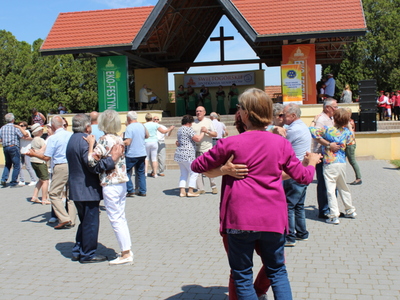 Na zdjęciu tańczący seniorzy. W tle scena na której występuje zespół śpiewaczy w zielonych kostiumach.