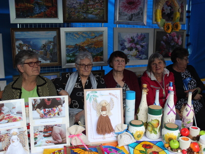 3)	Zdjęcie przedstawia pięć seniorek siedzących za ekspozycją rękodzieła Domu Dziennego Pobytu