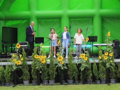 5)	Na zdjęciu znajdują się trzy dziewczynki występujące na scenie. Po lewej stronie mężczyzna w garniturze.