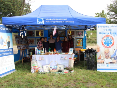 Zdjęcie wykonane w parku przy ulicy 11 Listopada w Sompolnie. Przedstawia pracownice Miejsko – Gminnego Ośrodka Pomocy Społecznej w Sompolnie stojące pod firmowym namiotem. W namiocie znajduje się wystawa prac uczestników Domu Dziennego Pobytu w Sompolnie.