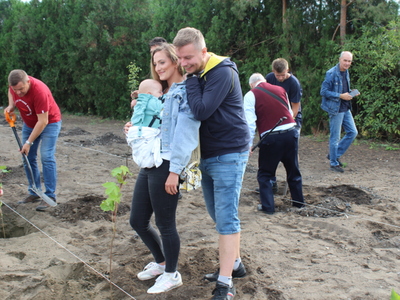 Na fotografii para z dzieckiem pozująca do zdjęcia. Przed nimi nowo posadzone drzewo. W tle osoby sadzące kolejne drzewa.