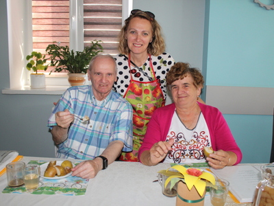 Zdjęcie ukazuje instruktorkę zajęć kulinarnych pozującą do zdjęcia z seniorami delektującymi się pieczonymi jabłkami.