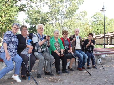 Na fotografii grupa seniorów siedząca na ławce w parku w piękny, słoneczny dzień.